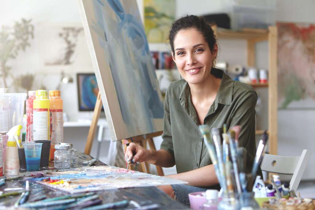Portrait of gorgeous excited young brunette female artist in casual blouse of khaki color, mixing oil paint on palette using painting knife, passionate about her occupation and process of creation